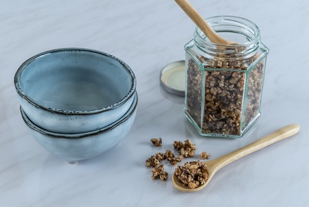 Homemade healthy and nutritious breakfast granola in a glass jar with wood spoon and blue ceramic bowls on marble kitchen table in fresh morning lightの写真素材
