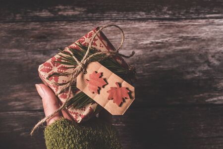 Hand holding a small secret Christmas present with creative handmade decorative rustic diy gift wrapped in red retro wrapping paper with natural vintage twine and spruce twigs as decor over old wood background giving it to someone special.の写真素材
