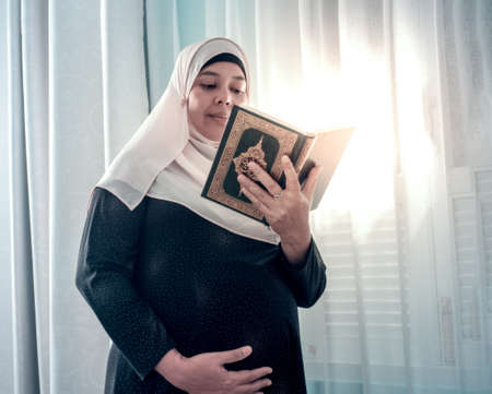 pregnant muslim woman with full traditional dress reading a book on her hand with smile face. Maternity  or motherhood concept.の写真素材