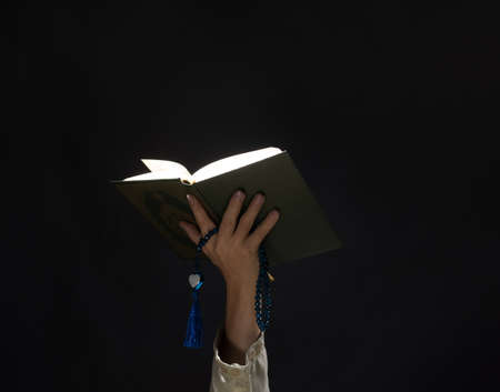 A man with his hand hold the Quran or Kuran ( the islamic holy book) on black background with rosary bead in ramadan night, muslim style and ramadan conceptの写真素材