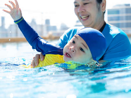 asian kid in a swimming suit excited on outdoor swimming pool over city buildings background with his motherの写真素材