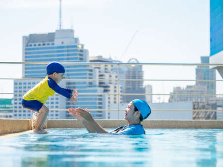 asian kid in a swimming suit excited on outdoor swimming pool over city buildings background with his motherの写真素材