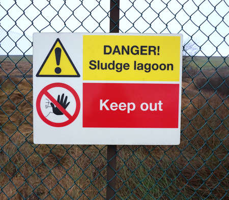 Yellow and red 'Danger Sludge Lagoon' Keep Out sign on a metal wire fenceの写真素材