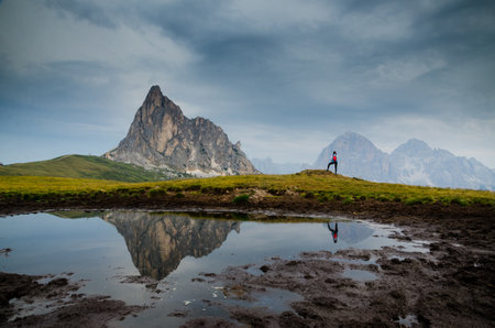 Young woman standing at a lake with reflecting a rocky mountain peak and a dark cloudy storm sky. Passo Giau before heavy rain.の写真素材