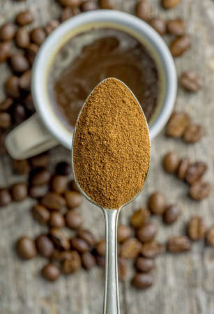 A close up of a spoonful of instant coffee powder over a cup of coffee and beansの写真素材