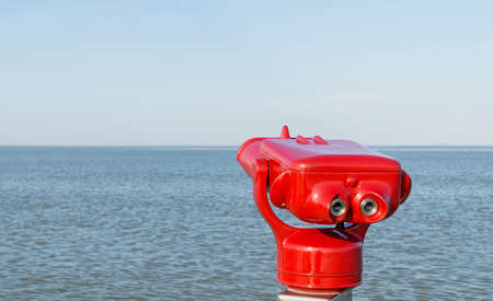 A set of red coin operated binoculars on the pier facing a blue sea and sky background with copy...