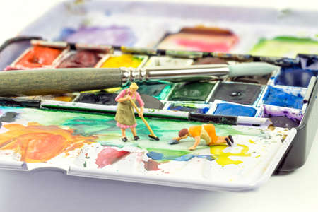 Two miniature ladies cleaning a messy artist's watercolor paint paletteの写真素材