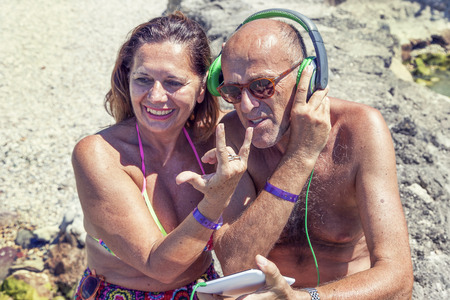 modern mature couple listening to music with headphones sitting on a pier. Concept of beautiful people having fun in summertimeの写真素材