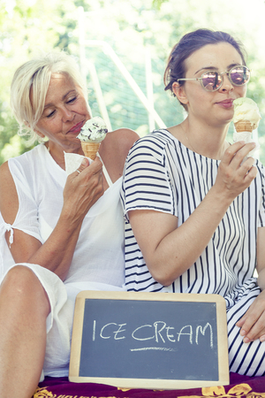 Modern mom and young daughter eating ice cream sitting on a deckchair in a water park on summertime. Concept of beautiful people having fun in summertimeの写真素材