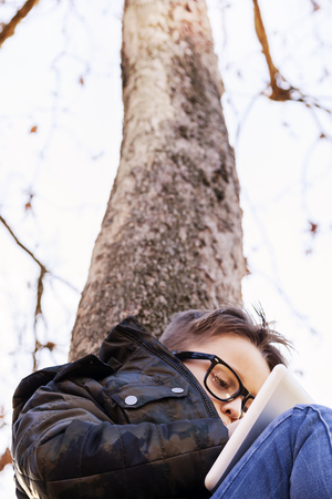 Elegant child plays with a tablet pc sitting on the branches of a tree in winter outdoorsの写真素材