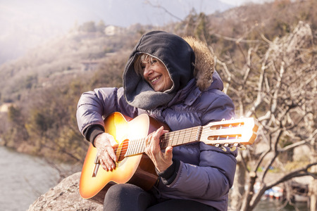 beautiful mature woman playing a guitar sitting on a rock in the mountains in a winter dayの写真素材