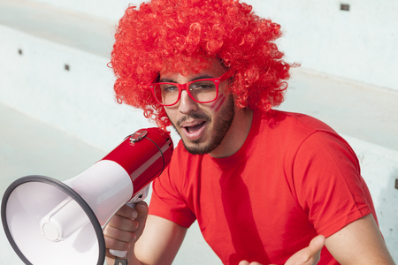 young supporter fan dressed in red color shouting in the megaphone to incite the team in the stands of a stadiumの写真素材
