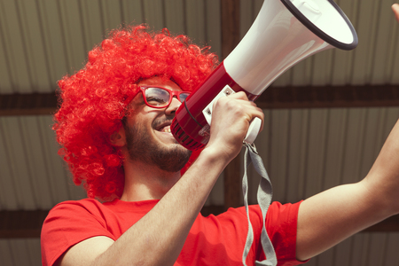 Young supporter fan dressed in red color shouting in the megaphone to incite the team in the stands of a stadiumの写真素材