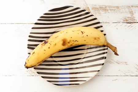 a ripe banana in a striped plate over a white wooden backgroundの写真素材