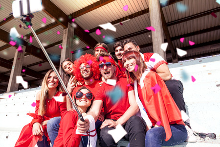 group of fans dressed in red color takes a selfie in the stands of a stadium during a sport eventの写真素材