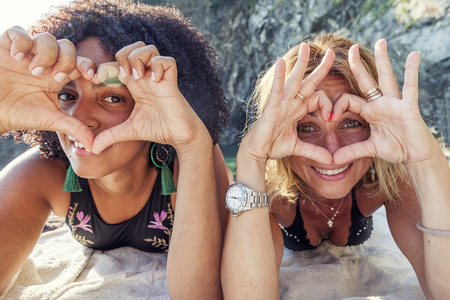 couple of beautiful women of different ethnicity showing heart shape with hands on beach vacationの写真素材