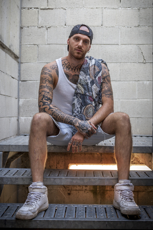 young caucasian tattooed singer rap posing sitting on the steps of a metal ladderの写真素材