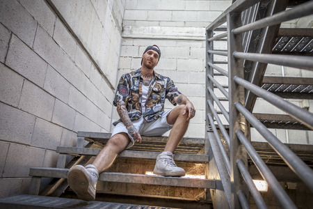 young caucasian tattooed singer rap posing sitting on the steps of a metal ladderの写真素材
