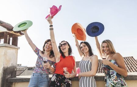 group of female friends having fun while drinking red wine on the roofs in summer throwing colorful hats - celebration and holiday conceptの写真素材