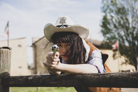 Portrait of a beautiful Chinese female cowgirl shooting with a weapon in the wild west behind a wooden fenceの写真素材