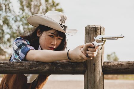 Portrait of a beautiful Chinese female cowgirl shooting with a weapon in the wild west behind a wooden fenceの写真素材
