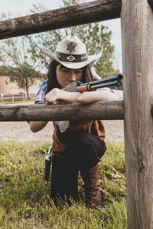 Portrait of a beautiful Chinese female cowgirl shooting with a weapon in the wild west behind a wooden fenceの写真素材