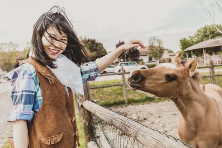 Portrait of a beautiful Chinese female cowgirl while stroking a foal in her fenceの写真素材