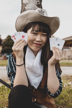 Portrait of a beautiful Chinese female cowgirl while playing with poker cards sitting outdoorsの写真素材
