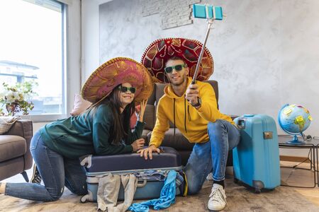 young couple sitting on the floor at home takes a selfie with smartphone wearing colorful sombrero...