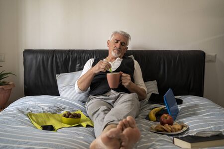 mature man is relaxing having breakfast in the bed of his home during tough quarantine at the time of coronavirusの写真素材