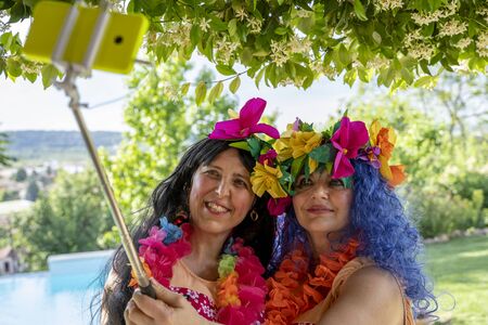 two smiling female friends dressed in Hawaiian costumes take a selfie with a smartphone by the poolの写真素材