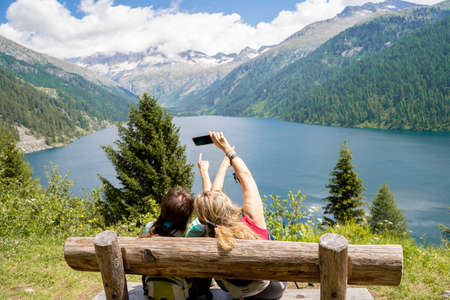 two female friends are photographing the landscape sitting on a wooden bench in front of a mountain lakeの写真素材