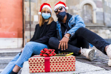 young multi-ethnic couple exchanges Christmas gifts wearing protective masks during the coronavirus...