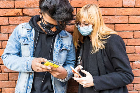 young multiethnic couple with protective masks in front of brick wall using smartphones to share...