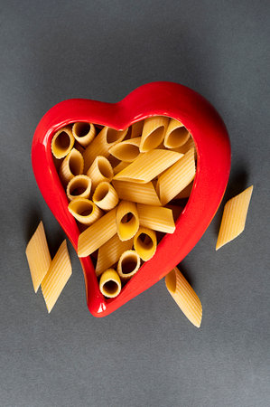 fresh pasta penne rigate in a red heart shaped container on a gray background. zenith view