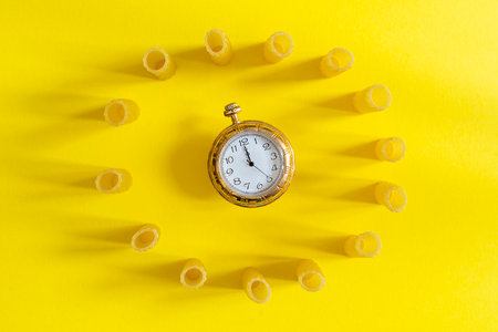 small group of fresh macaroni rigati lined up in a circle with a clock in the middle on yellow background. concept of italian cuisineの写真素材