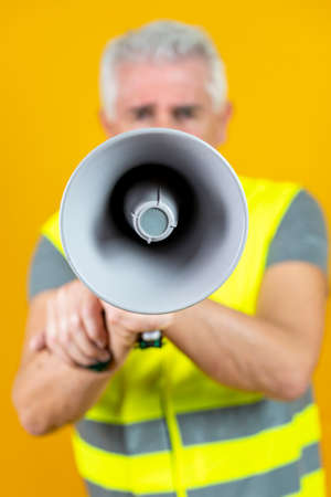 mature worker wearing reflective vest screaming into megaphone isolated on yellow backgroundの写真素材