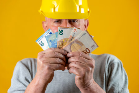 worker with yellow helmet showing euro banknotes in the foreground on yellow backgroundの写真素材