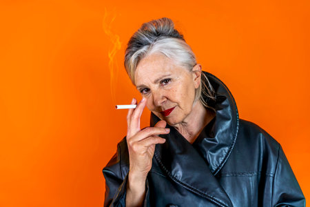 Funny portrait of mature woman. Sophisticated lady having fun dressed in a leather coat smoking a cigarette. Mature woman on colored backgroundsの写真素材