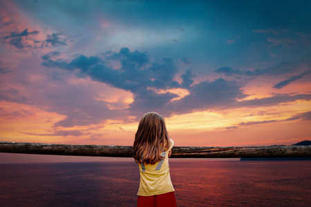 little cute girl seen from behind leaning against a wooden fence is looking at stunning sunset on the seaの写真素材
