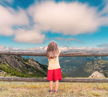 little cute girl seen from behind leaning against a wooden fence is looking at the mountains and cloudsの写真素材