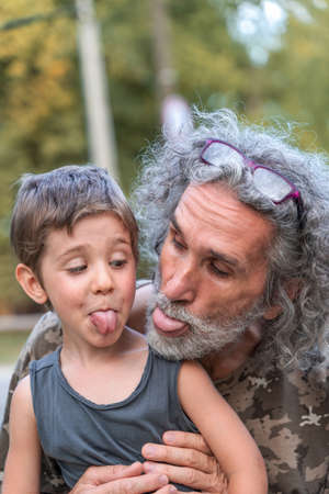 portrait of father with long gray hair and son are showing the tongue outdoorsの写真素材