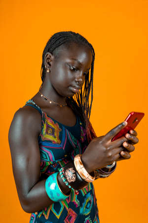 young african female with traditional dress and bracelets is chatting with a smartphone. Studio shot on yellow backgroundの写真素材