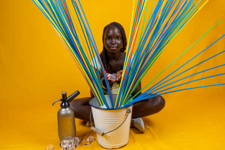 young cute african teen sitting preparing a giant cocktail with ice and long colorful drinking straws. Studio shot on yellow backgroundの写真素材