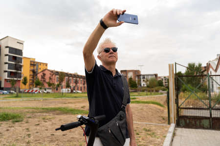 commuter hipster man with electric scooter take a selfie with smartphone in the suburbs. Eco transport conceptの写真素材