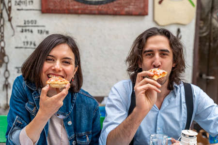 smiling beautiful couple eating pizza outdoors looking at cameraの写真素材