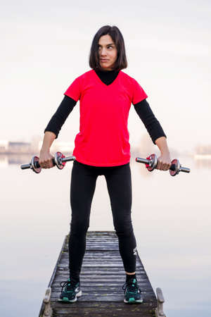 attractive young woman is doing exercises with dumbbells on a harbor quay in winter - health and...