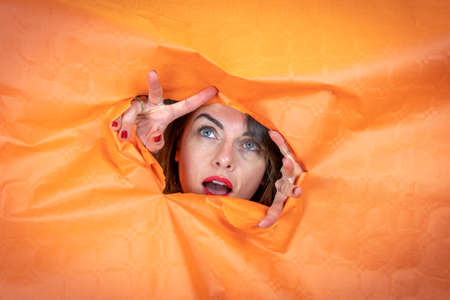 portrait of attractive young woman is coming out of a hole in a sheet of orange paperの写真素材