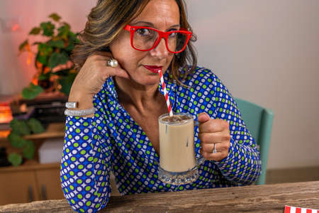 woman dressed in blue shirt and red glasses sitting drinking cold coffee through a strawの写真素材