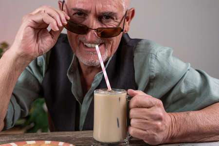 casual dressed mature man with sunglasses sitting at a table drinking iced coffee through a strawの写真素材